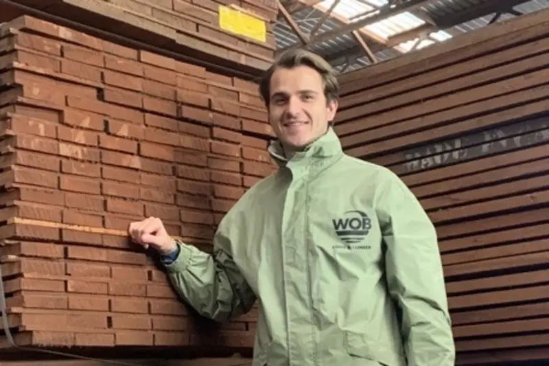 Geschäftsführer von WOB Timber in grüner Arbeitsjacke lächelnd vor gestapeltem Schnittholz im Lager.